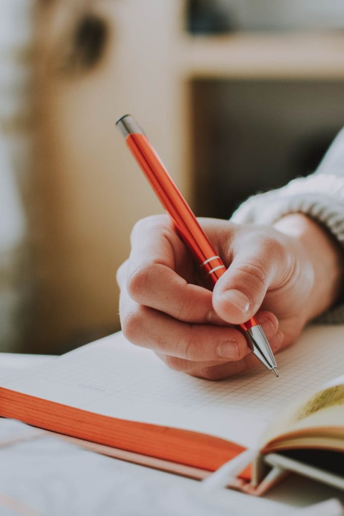 A person writing in a notebook with a red ballpoint pen, showcasing detail and focus.